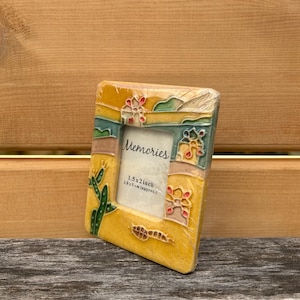 May include: A rectangular ceramic picture frame with a yellow base, featuring raised relief designs of green cacti and colorful desert-inspired motifs. The center insert displays the text "Memories" and "1.5 x 2 inch". The frame is wrapped in clear plastic.