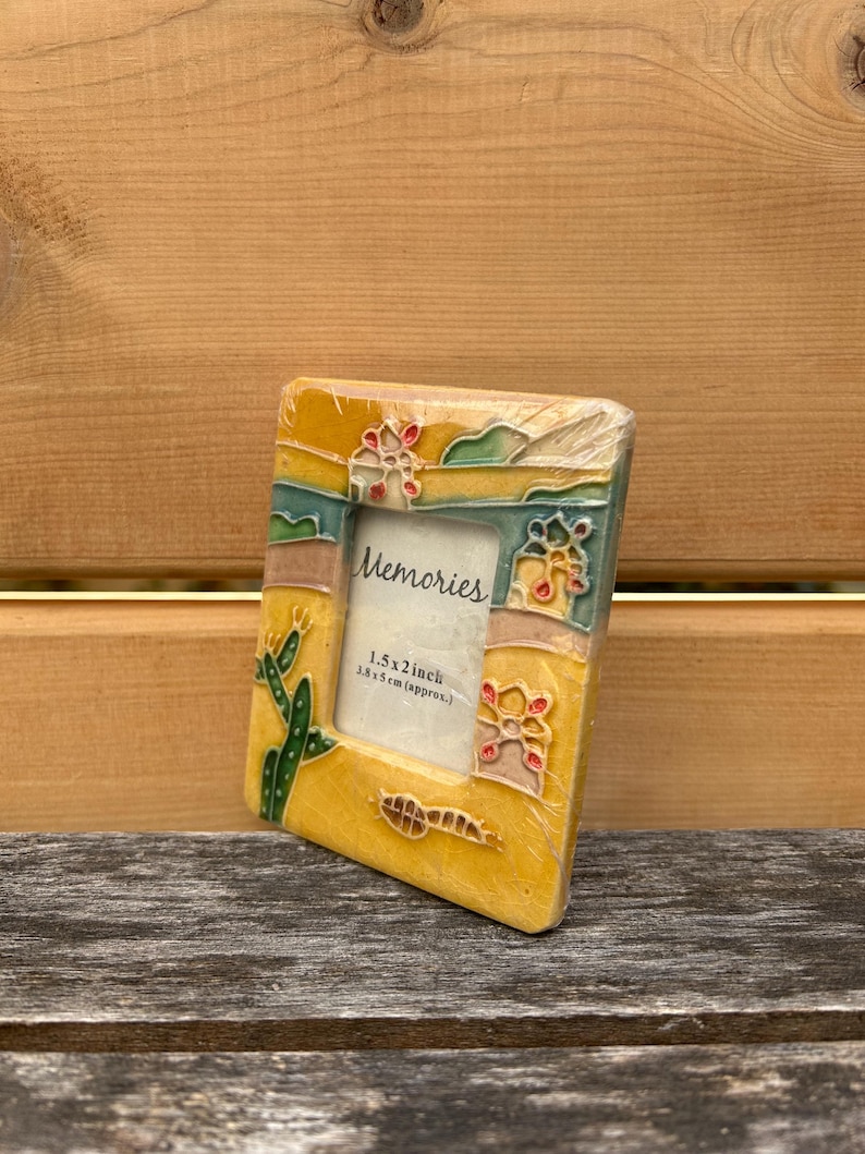 May include: A rectangular ceramic picture frame with a yellow base, featuring raised relief designs of green cacti and colorful desert-inspired motifs. The center insert displays the text "Memories" and "1.5 x 2 inch". The frame is wrapped in clear plastic.