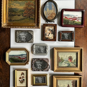 May include: A collection of framed art pieces, including landscape paintings, floral arrangements, and a vintage airplane. Frames vary in shape and color, with gold, silver, and brown tones. The artwork is displayed on a white background against a dark wood surface.