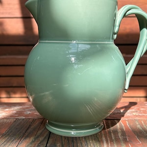 May include: A sage green ceramic pitcher with a rounded body, a flared rim featuring a pouring spout, and a curved handle attached to the side. The surface has a smooth, glossy finish with a raised horizontal band encircling the upper section.