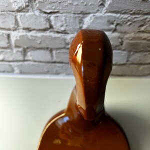 May include: A glossy, brown ceramic sculpture with a smooth, curved form. The sculpture has a rounded top and a wider base, with a reflective surface that catches the light. The piece is set against a neutral background.