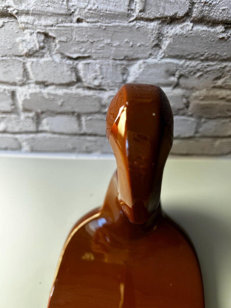 May include: A glossy, brown ceramic sculpture with a smooth, curved form. The sculpture has a rounded top and a wider base, with a reflective surface that catches the light. The piece is set against a neutral background.