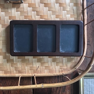 May include: A rectangular dark brown wooden frame featuring three vertical, side-by-side openings with black inserts. The frame has a smooth, solid construction with rounded corners, designed to hold three separate items within its distinct sections.
