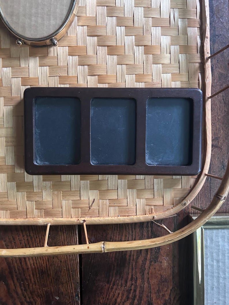 May include: A rectangular dark brown wooden frame featuring three vertical, side-by-side openings with black inserts. The frame has a smooth, solid construction with rounded corners, designed to hold three separate items within its distinct sections.