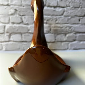 May include: A brown ceramic swan-shaped decorative bowl. The bowl has a glossy finish and a curved design. The swan's neck extends upwards, and the bowl's base is wide and open. The background is a white brick wall.