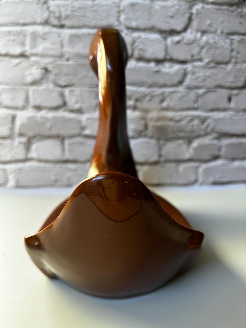 May include: A brown ceramic swan-shaped decorative bowl. The bowl has a glossy finish and a curved design. The swan's neck extends upwards, and the bowl's base is wide and open. The background is a white brick wall.