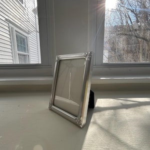 May include: A rectangular silver-toned picture frame with a smooth, metallic finish and decorative corner detailing. The frame features a clean, neutral-colored matting inside and is supported by a black easel-style stand on the back for display.