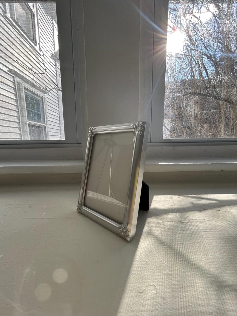 May include: A rectangular silver-toned picture frame with a smooth, metallic finish and decorative corner detailing. The frame features a clean, neutral-colored matting inside and is supported by a black easel-style stand on the back for display.