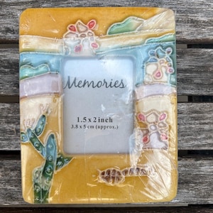 May include: A rectangular yellow picture frame with a raised desert-themed relief featuring a green cactus, pink flowers, and blue accents. The center insert displays the word Memories and the text 1.5 x 2 inch 3.8 x 5 cm (approx.).