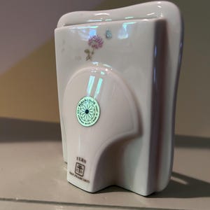 May include: A white ceramic picture frame with a glossy finish, featuring a small floral decal of a pink flower and a blue butterfly. A circular green sticker is centered on the back, and the text FERN SAN FRANCISCO is printed near the base.