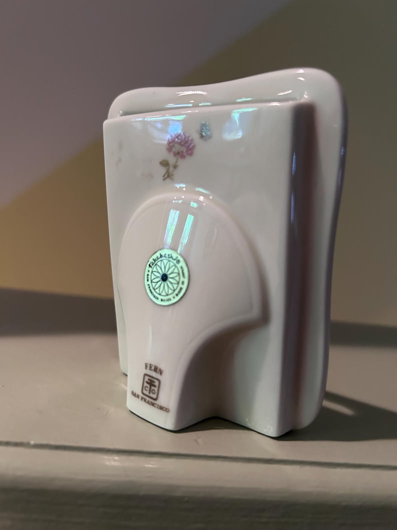 May include: A white ceramic picture frame with a glossy finish, featuring a small floral decal of a pink flower and a blue butterfly. A circular green sticker is centered on the back, and the text FERN SAN FRANCISCO is printed near the base.