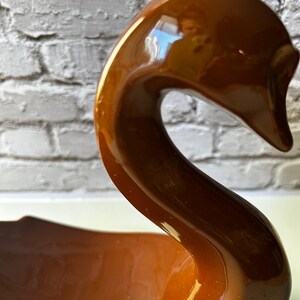 May include: A glossy, brown ceramic swan figurine with a long, curved neck and a wide base. The swan has a smooth, reflective surface and is set against a white brick wall. The figurine is a decorative item.