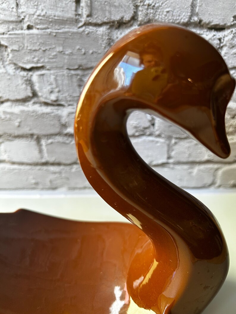 May include: A glossy, brown ceramic swan figurine with a long, curved neck and a wide base. The swan has a smooth, reflective surface and is set against a white brick wall. The figurine is a decorative item.