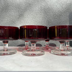 May include: A set of five matching dessert bowls made of deep red glass. Each bowl features a wide, circular basin supported by a short, clear glass stem and a sturdy, flat circular base, creating a uniform look for serving small portions.