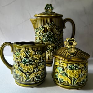 May include: A three-piece ceramic coffee set in shades of olive green and yellow. The set includes a coffee pot, creamer, and sugar bowl, all decorated with a floral pattern. The coffee pot has a lid and handle.