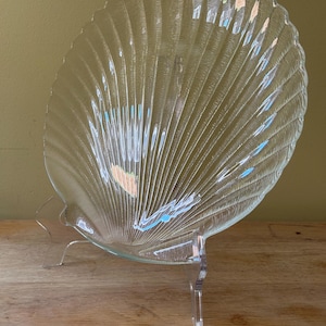 May include: A clear glass seashell-shaped serving dish with a textured surface, displayed on a clear plastic stand. The dish has a scalloped edge and radial lines, creating a fan-like pattern. The stand is clear and has a star-shaped base.