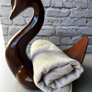 May include: A brown ceramic swan-shaped holder with a rolled-up beige towel. The swan has a glossy finish and a curved neck. The towel is made of a textured fabric.