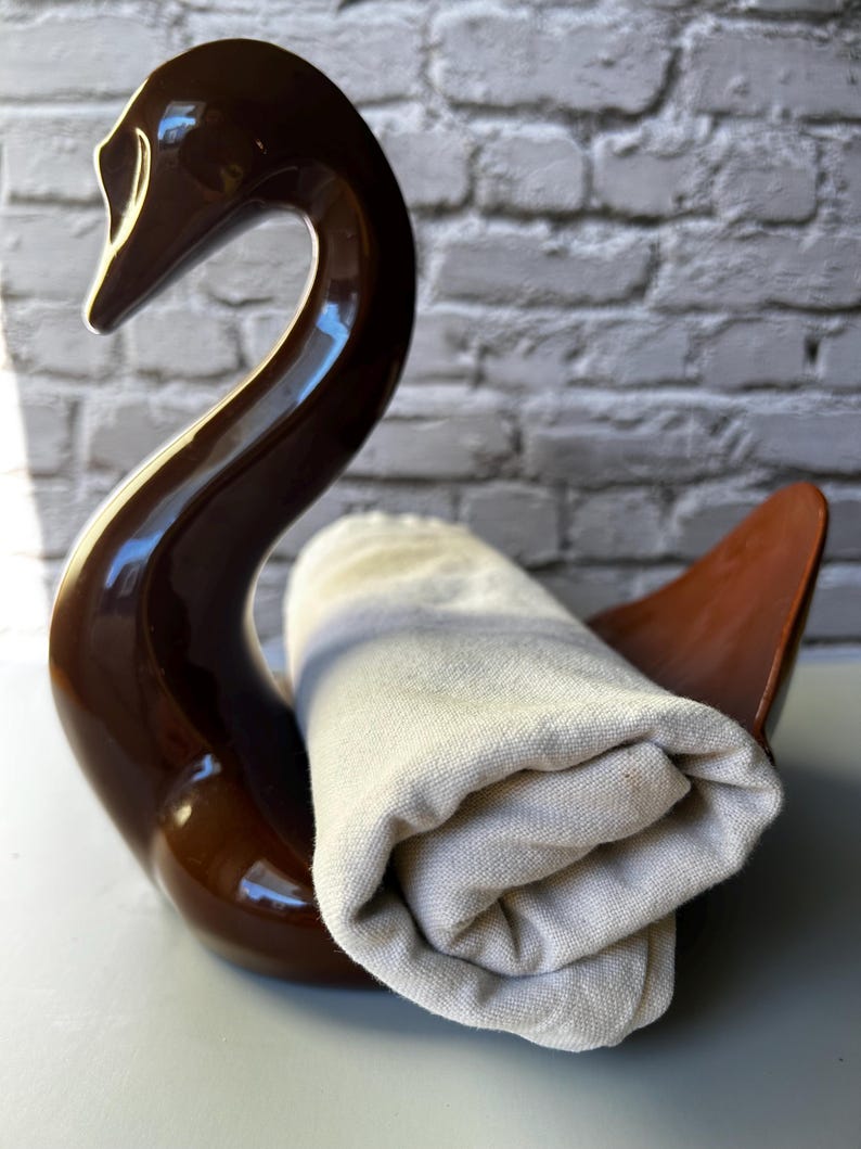 May include: A brown ceramic swan-shaped holder with a rolled-up beige towel. The swan has a glossy finish and a curved neck. The towel is made of a textured fabric.