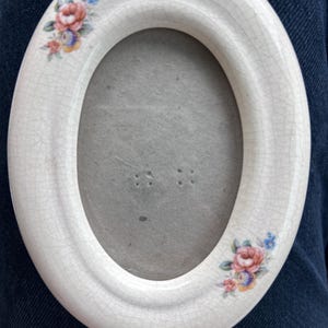 May include: An oval ceramic picture frame with a cream-colored crackle glaze finish. The frame features small, hand-painted floral clusters in pink, blue, and yellow at the top left and bottom right corners, surrounding a plain gray center.