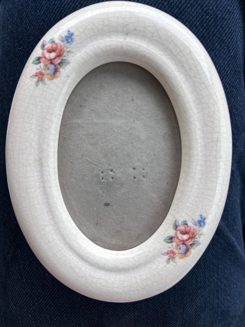 May include: An oval ceramic picture frame with a cream-colored crackle glaze finish. The frame features small, hand-painted floral clusters in pink, blue, and yellow at the top left and bottom right corners, surrounding a plain gray center.