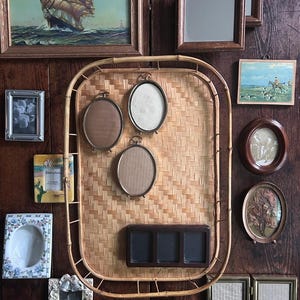 May include: A large rectangular bamboo tray mounted on a dark wood wall, serving as a display for three oval metal frames and a dark brown rectangular triple-frame. The tray is surrounded by various wall-mounted picture frames of different shapes.