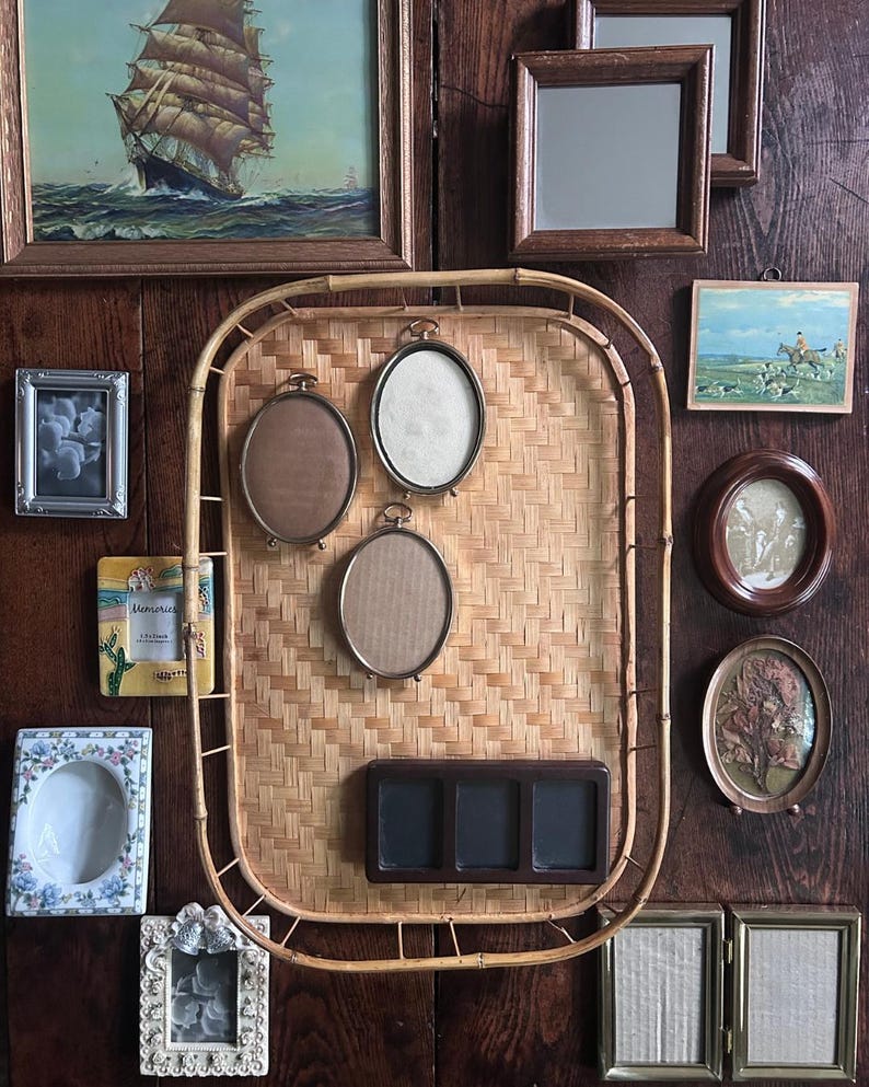 May include: A large rectangular bamboo tray mounted on a dark wood wall, serving as a display for three oval metal frames and a dark brown rectangular triple-frame. The tray is surrounded by various wall-mounted picture frames of different shapes.