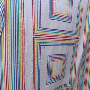 May include: A white fabric textile featuring a repeating pattern of concentric rainbow-colored squares and vertical stripes. The design includes thin lines in red, orange, yellow, green, blue, and purple. The signature "Vera" is printed in script.
