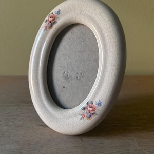 May include: An oval ceramic picture frame with a cream-colored crackle glaze finish. The frame features two small floral decals with pink, blue, and orange flowers positioned at the top left and bottom right corners of the smooth, rounded border.