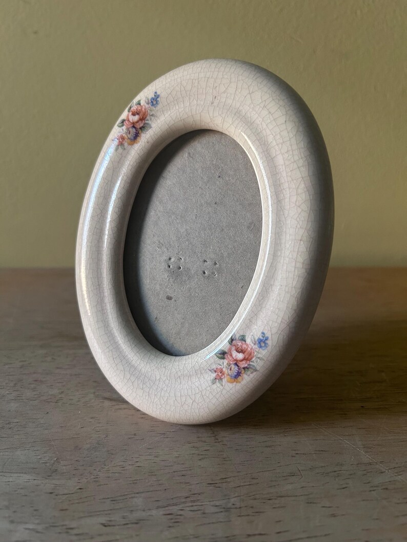 May include: An oval ceramic picture frame with a cream-colored crackle glaze finish. The frame features two small floral decals with pink, blue, and orange flowers positioned at the top left and bottom right corners of the smooth, rounded border.