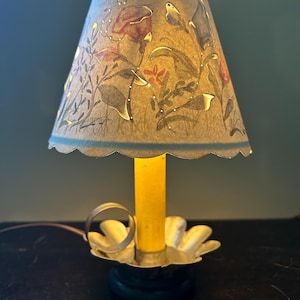 May include: A small table lamp featuring a yellow candle-shaped bulb and a scalloped metal base with a finger loop. The conical paper shade is decorated with a painted floral pattern in red, blue, and green, featuring cut-out details.