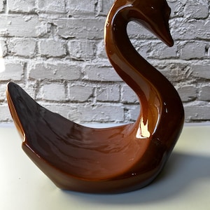 May include: A brown ceramic swan figurine with a glossy finish. The swan has a long, curved neck and a wide, open base, creating a decorative bowl shape. The background features a white brick wall.