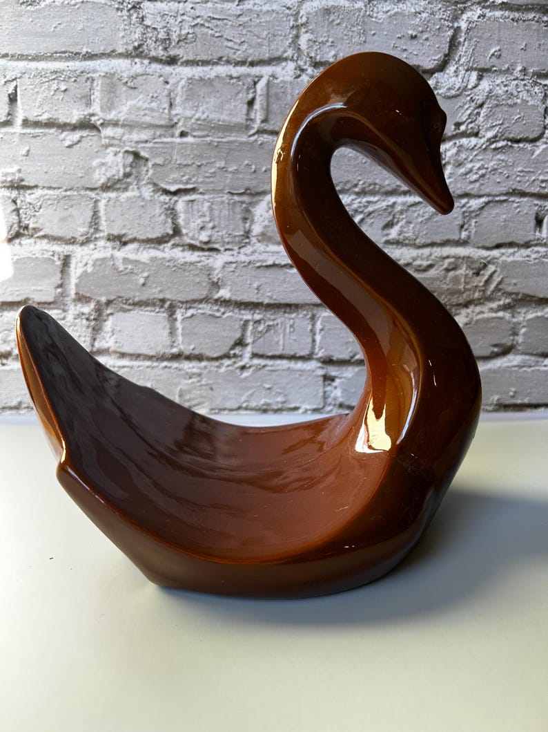 May include: A brown ceramic swan figurine with a glossy finish. The swan has a long, curved neck and a wide, open base, creating a decorative bowl shape. The background features a white brick wall.