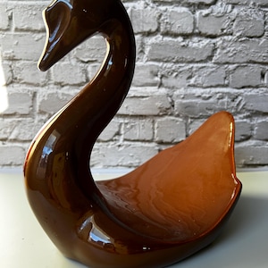 May include: A brown ceramic swan figurine with a glossy finish. The swan has a curved neck and a wide, open body, creating a decorative piece. The figurine is likely a decorative item for a home or office.
