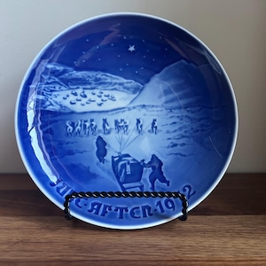 May include: A round porcelain collector plate in shades of blue and white featuring a winter scene of a dog sled team traveling across a snowy landscape toward a distant village under a starry sky, with the text "AFTER 1992" printed at the bottom.