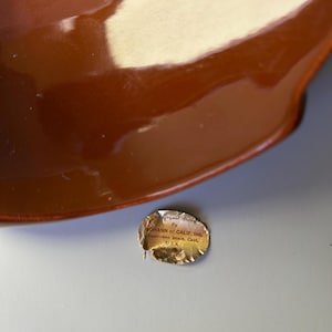 May include: A glossy, brown ceramic bowl with a curved rim. The bowl has a reflective surface. A small, torn sticker with text is visible near the bowl. The sticker reads "JOHANN of CALIF. Inc. Capistrano Beach, Calif. U.S.A."