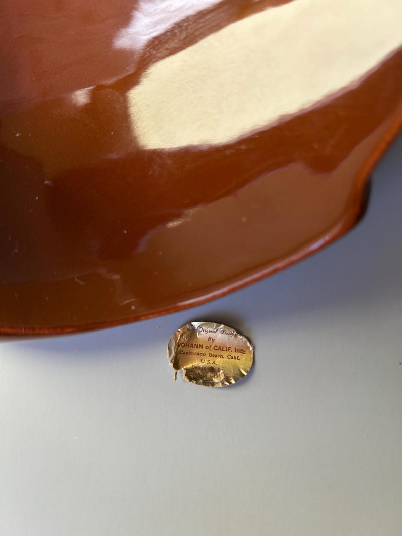 May include: A glossy, brown ceramic bowl with a curved rim. The bowl has a reflective surface. A small, torn sticker with text is visible near the bowl. The sticker reads "JOHANN of CALIF. Inc. Capistrano Beach, Calif. U.S.A."