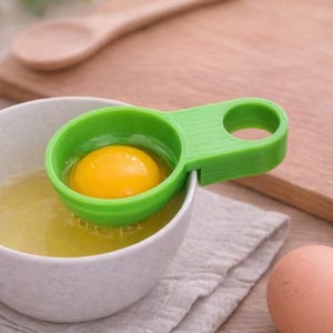 Può includere: Un separatore di uova verde tiene un tuorlo d'uovo giallo brillante sopra una ciotola bianca piena di albumi. Due uova marroni sono a destra. Un cucchiaio di legno e un tagliere sono sullo sfondo. L'utensile da cucina è progettato per separare i bianchi dai tuorli.