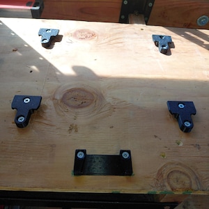 May include: A wooden surface with six black metal brackets attached with screws. The brackets are various shapes and sizes, and are mounted on a light-colored wood surface. The wood has visible grain and knots.