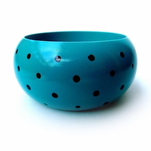 May include: A wide, rounded bangle bracelet featuring a vibrant turquoise blue base color with a repeating pattern of small, solid black polka dots distributed across the smooth, glossy surface of the circular accessory.