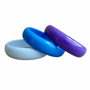 May include: Three smooth, rounded bangle bracelets arranged in a stack. The bracelets feature a glossy finish in distinct shades of light blue, vibrant medium blue, and deep purple. Each bangle has a solid, uniform color and a simple circular shape.