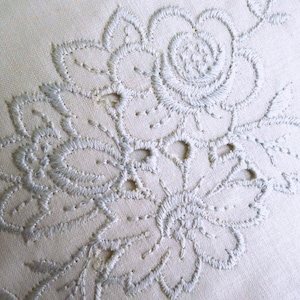 May include: Close-up of a cream-colored textile featuring intricate, light blue floral embroidery. The design includes a large rose and other stylized flowers with detailed stitching and openwork.