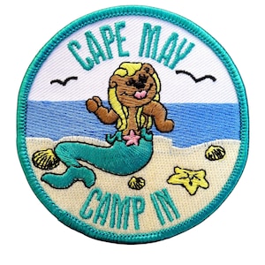May include: A circular embroidered patch featuring a brown mermaid with blonde hair on a sandy beach. The text reads CAPE MAY at the top and CAMP IN at the bottom in teal. The design includes a blue ocean, two black birds, and yellow seashells.
