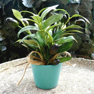 May include: A small artificial olive plant with dark green oval fruits and pointed green leaves in a solid teal ceramic pot. The base of the plant is decorated with a natural tan jute twine bow tied around the stems above the pot rim.