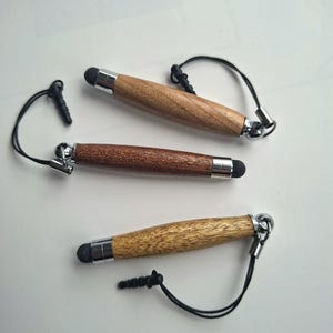 Könnte beinhalten: Drei Eingabestifte mit Holzgehäusen in verschiedenen hellen, mittleren und dunklen Brauntönen. Jeder Stift verfügt über eine silberne Metallspitze, eine schwarze Gummikontaktspitze und ein schwarzes Kordelband mit einem kleinen Stecker.