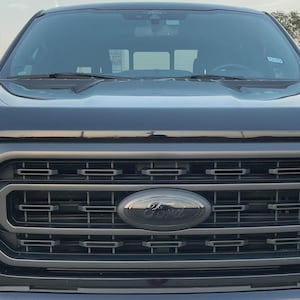 May include: Close-up of a black Ford truck grille. The grille features a black Ford emblem in the center, surrounded by a geometric pattern. The hood and windshield of the truck are visible above the grille.