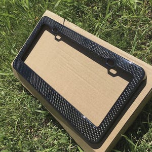 May include: A black license plate frame with a carbon fiber weave pattern. The frame has a glossy finish and is rectangular with rounded corners. It is sitting on a cardboard box, with green grass in the background.