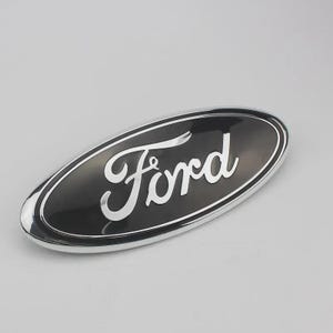 May include: Oval-shaped black and silver Ford emblem. The word "Ford" is written in a cursive font in silver. The emblem has a silver border and is set against a light gray background.