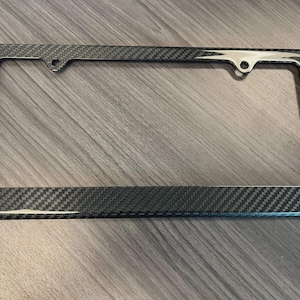 May include: A rectangular license plate frame with a carbon fiber weave pattern. The frame has a polished chrome accent on the top edge and around the mounting holes. The frame is designed to fit a standard U.S. license plate.