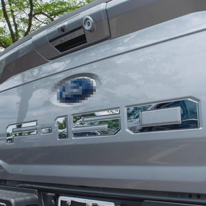 May include: Close-up of the rear of a silver Ford F-150 truck. The tailgate features the Ford emblem and the model name in chrome lettering. The truck is parked outdoors.