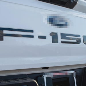 May include: Close-up of the rear of a white Ford F-150 truck. The black lettering of "F-150" is prominently displayed on the tailgate. The Ford logo is visible above the lettering.